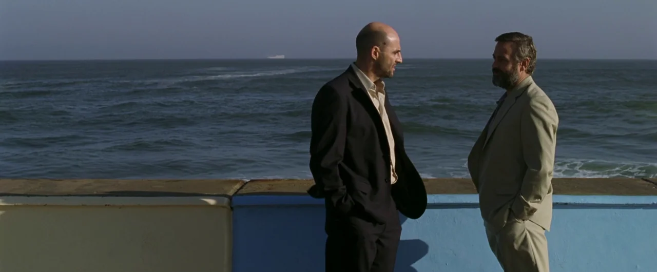Syriana (2005) movie still: fullbody — Two men stand on a concrete seawall bordering a vast ocean under clear skies. They are engaged in a quiet…