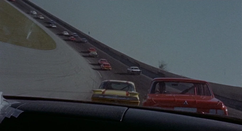 Vanishing Point (1971) movie still: wide — A steeply banked asphalt racetrack stretches upward into a pale sky under daylight. A pack of stock cars…