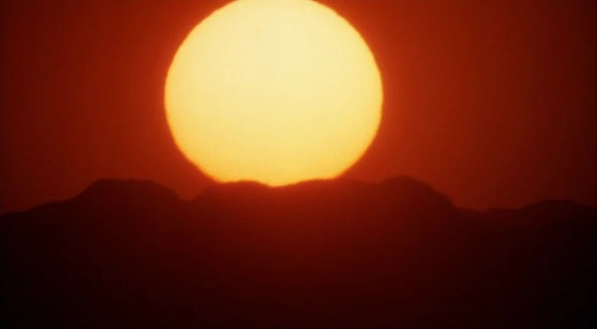 Dust Devil (1992) movie still: establishing — A large, glowing orange sun dips behind a dark, jagged mountain horizon line. The scene is dominated…