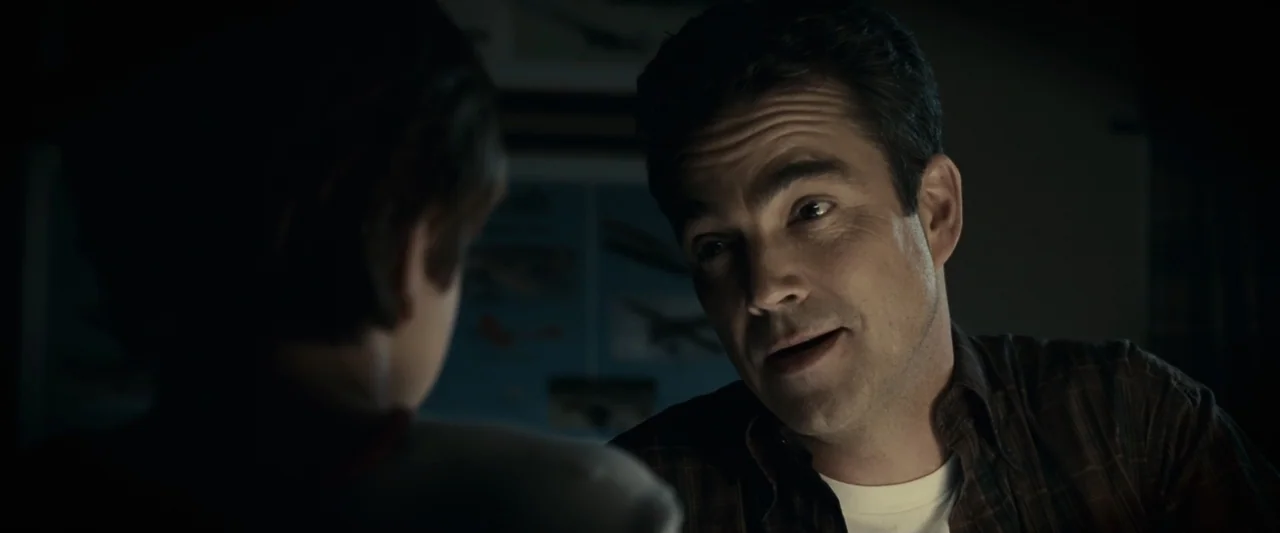 Green Lantern (2011) movie still: medium — A dimly lit room contains posters on the wall in the blurred background. A man leans toward a child while…