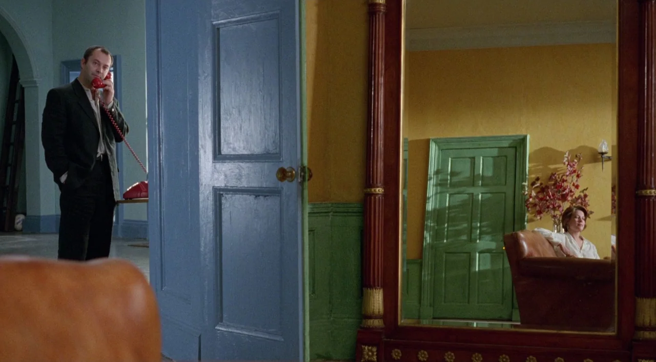 Shallow Grave (1994) movie still: medium — A hallway features bright yellow walls and a large, ornate floor mirror. A man stands in the foreground…
