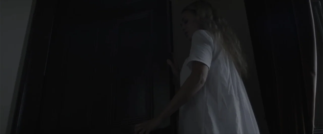 Cinderella's Curse (2024) movie still: medium — A dimly lit interior shows a dark wooden door partially open in a room. A woman wearing a white…