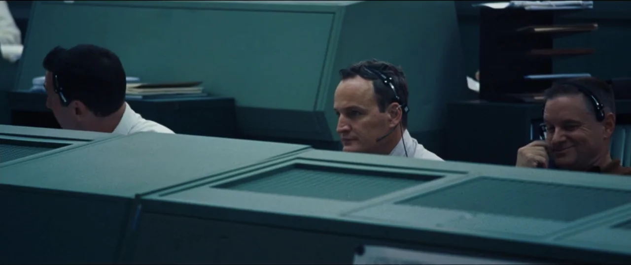 First Man (2018) movie still: medium — A 1960s era mission control room features rows of green metal consoles and monitors. Three men in headsets…
