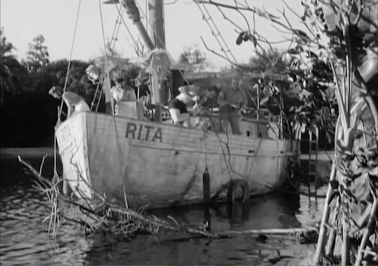 Creature from the Black Lagoon (1954) movie still: wide — A wooden fishing boat named Rita is docked in a calm, tree-lined river. Several…