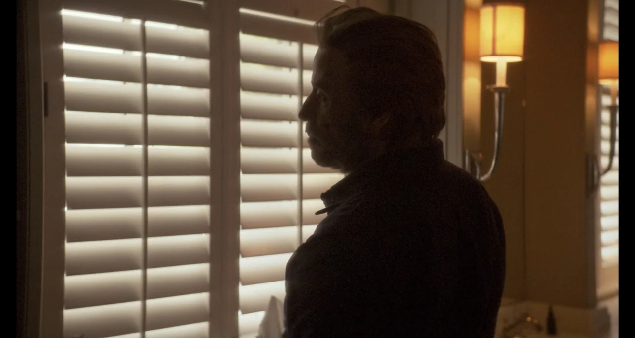 The Poison Rose (2019) movie still: medium — A dimly lit room features a window covered with white horizontal plantation shutters. A man stands in…