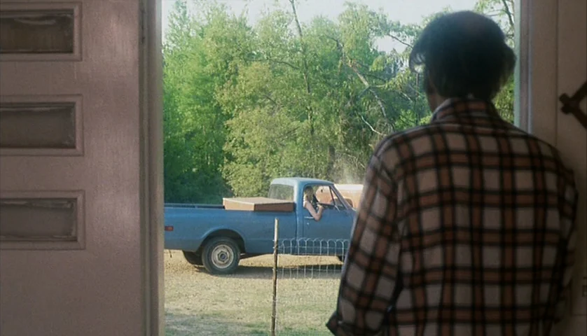 Cockfighter (1974) movie still: medium — A man in a plaid shirt stands in a doorway looking at a truck. A blue pickup truck with a woman driver…