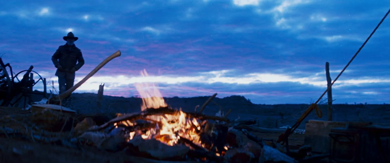 Razorback (1984) movie still: wide — A rugged campsite at dusk features a small campfire flickering against a vast landscape. A man wearing a cowboy…