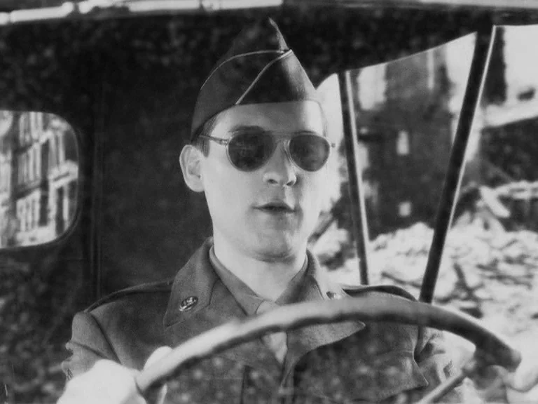 The Good German movie still: medium — A soldier sits inside a military jeep against a backdrop of ruined buildings. He holds the steering wheel…