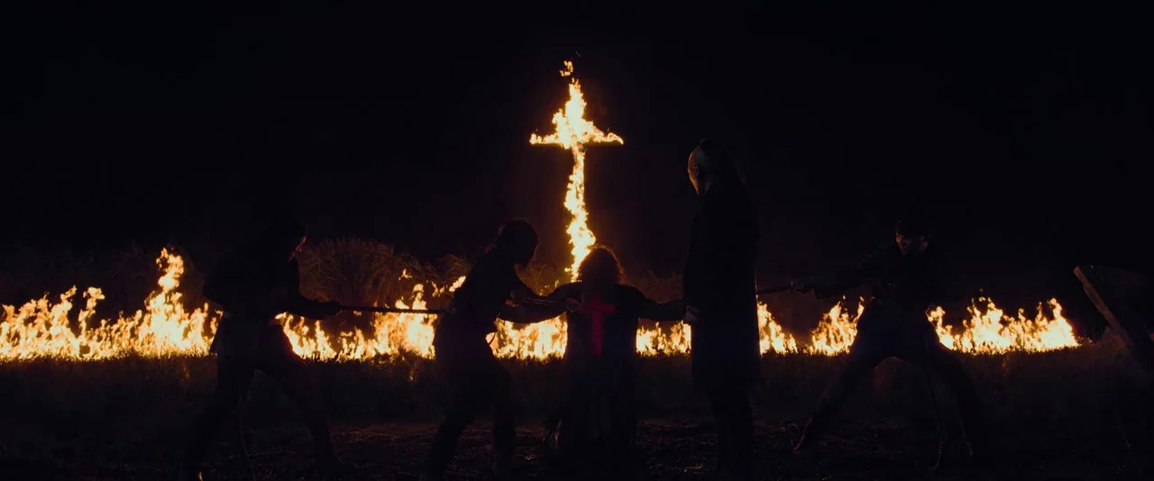 Apostle (2018) movie still: wide — An open dark field features a large flaming cross structure and fire-line backdrop. Four individuals in dark…