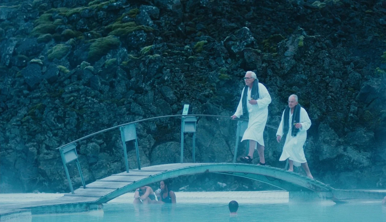 Land Ho! (2014) movie still: wide — A geothermal pool is surrounded by large dark mossy volcanic rock formations. Two elderly men in white bathrobes…