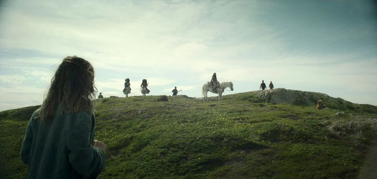 Peter Pan & Wendy (2023) movie still: wide — A grassy hill with rocky outcrops stands under a pale overcast sky. A group of figures and a white…