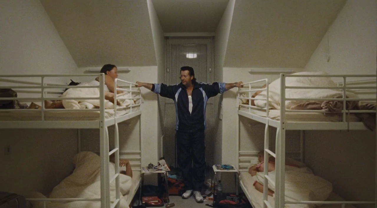 Paradise: Hope (2013) movie still: fullbody — A spartan dormitory with four white metal bunk beds and angled walls. A man in a blue tracksuit stands…