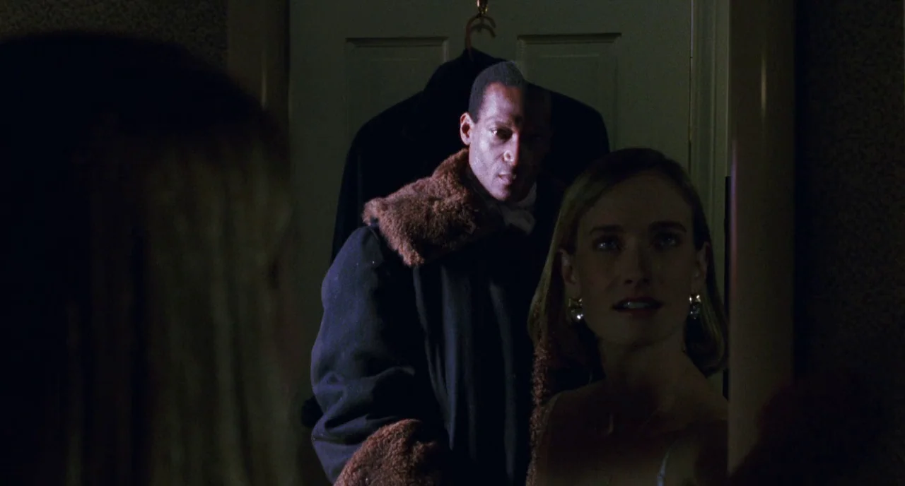 Candyman (1992) movie still: medium — A dark dressing room featuring a mirror reflecting two people standing together closely. A man stands behind a…