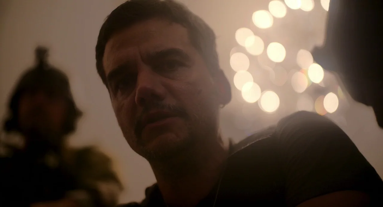Civil War (2024) movie still: closeup — A man looks down at the camera inside a dimly lit interior space. Soft out-of-focus lights appear in the…