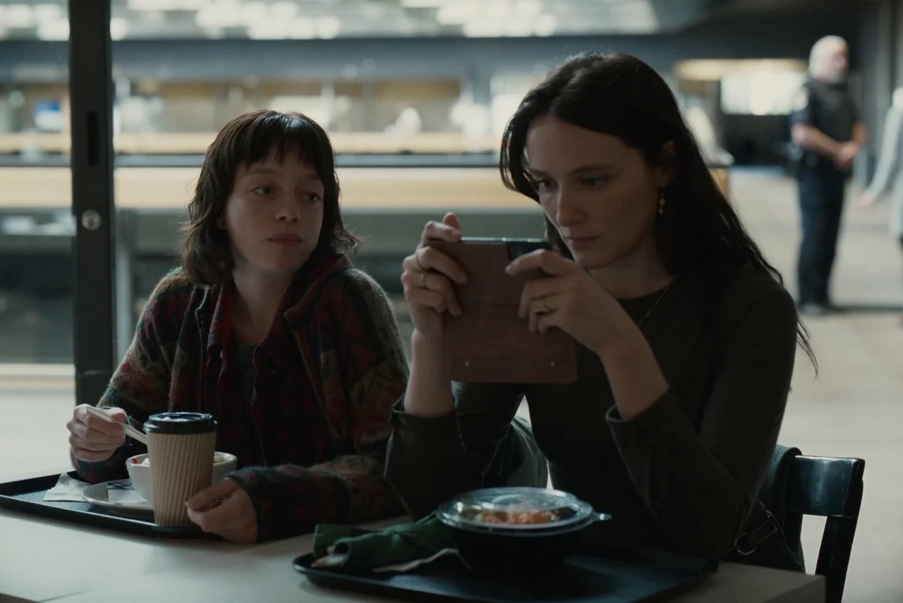 Red Rooms (2023) movie still: medium — Two women sit at a white table in a modern, sparsely decorated indoor facility. One woman uses a smartphone…