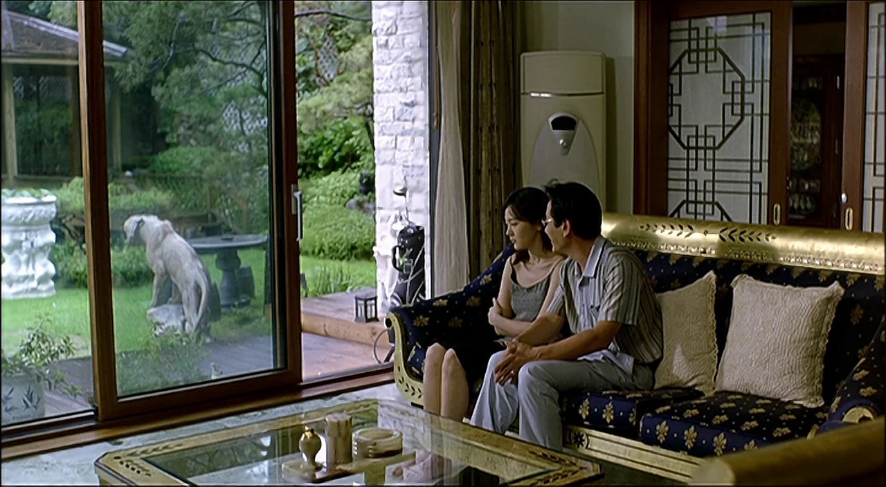 3-Iron (2004) movie still: medium — A spacious room features a low gold-trimmed sofa facing a large sliding glass door. The couple sits together on…