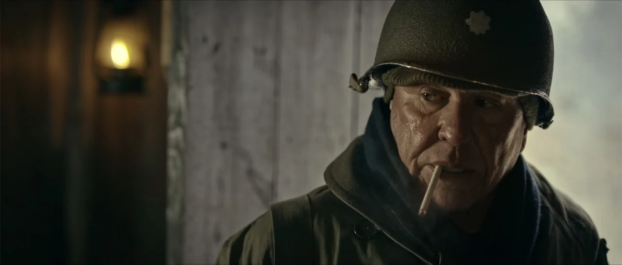 Battle of the Bulge (1965) movie still: closeup — A dimly lit room features wooden walls and a glowing lamp out of focus. An older soldier wearing a…