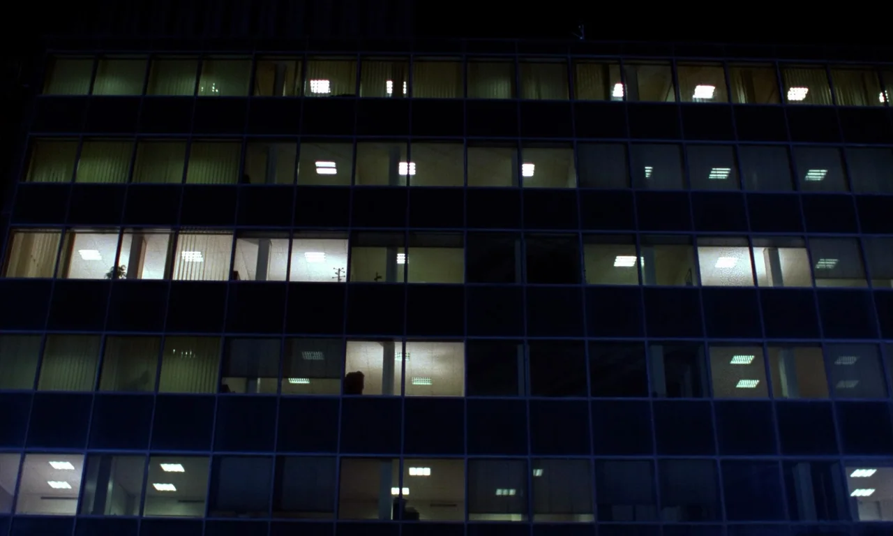 Demons 2 (1986) movie still: establishing — A modern commercial building facade at night displays a grid of illuminated office windows. The dark…