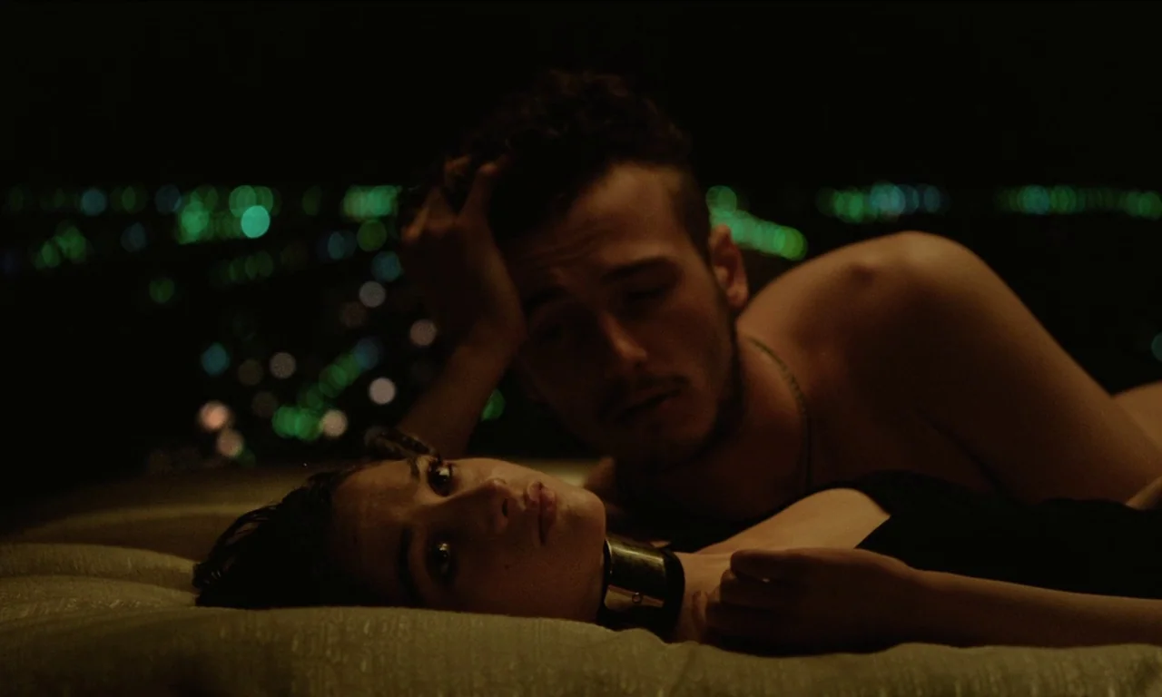 Vox Lux (2018) movie still: medium — A dark, blurred city skyline glows in the background behind a bed. A young man and woman lie on sheets in a…