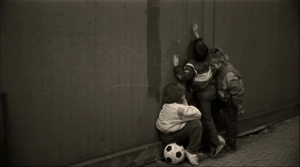 Wings Of Desire (1987) movie still: fullbody — Three children stand against a tall, featureless concrete wall in a narrow outdoor alleyway. One…