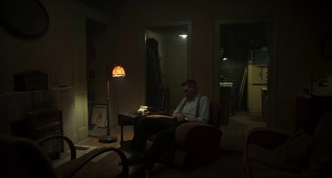 The Little Stranger (2018) movie still: wide — A dim 1950s interior space features period-accurate furniture, a vintage floor lamp, and doorway…