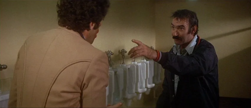 California Split (1974) movie still: medium — A public restroom with beige walls features a row of white porcelain urinals. A man in a dark jacket…