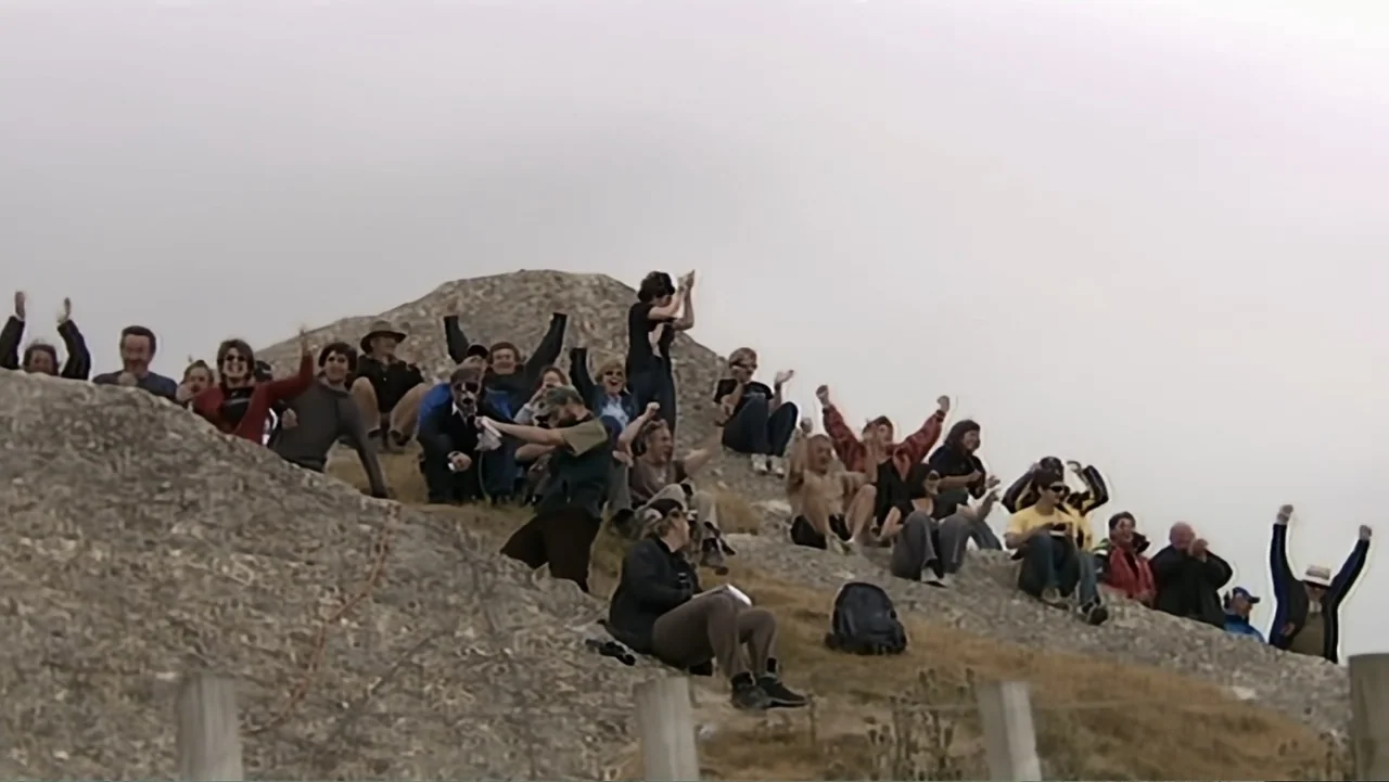 Wild Wild Space (2024) movie still: wide — A crowd of people sits on a rocky, dry hillside under a grey sky. The group waves and applauds with their…