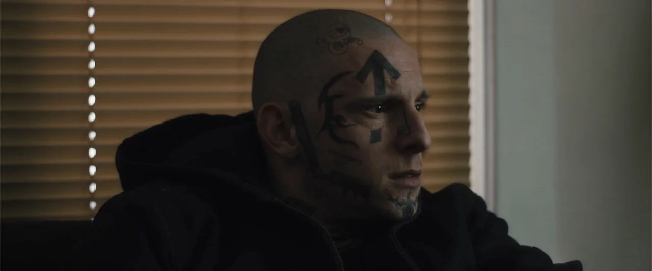 Skin (2009) movie still: closeup — A man sits in an indoor space with horizontal blinds visible in the background. The man displays prominent facial…