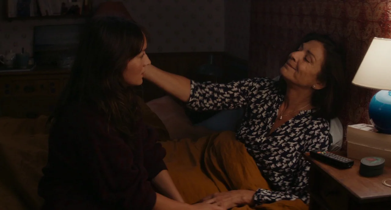 Anaïs in Love (2021) movie still: medium — A dimly lit room with patterned wallpaper features a woman sitting beside a bed. An older woman lies…