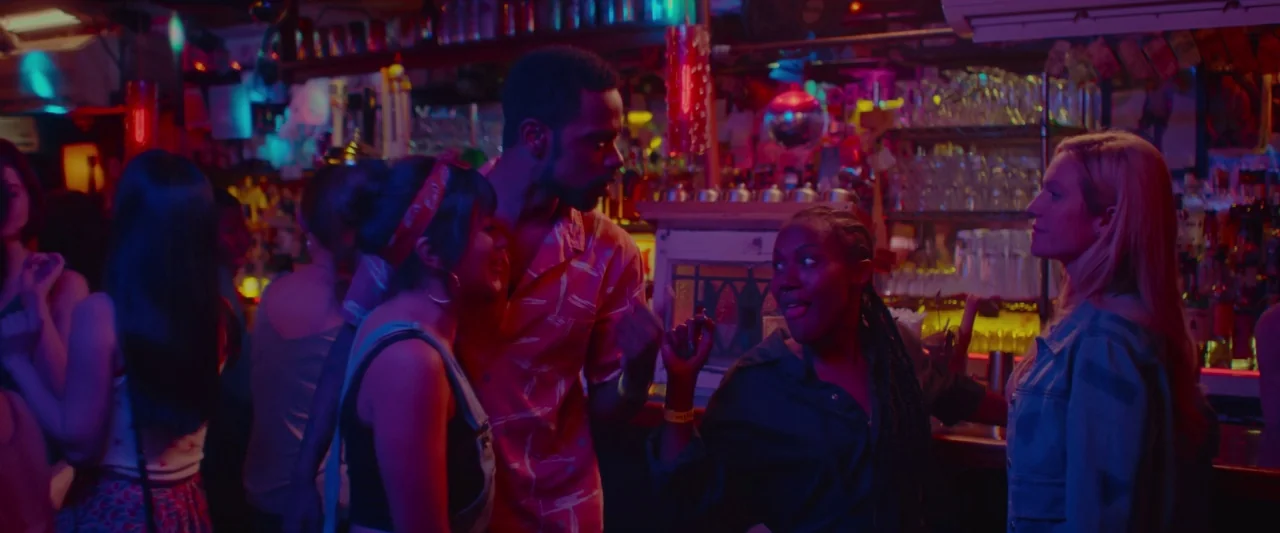 Someone Great (2019) movie still: medium — A crowded bar interior is lit by intense saturated neon light fixtures. A group of four young adults…