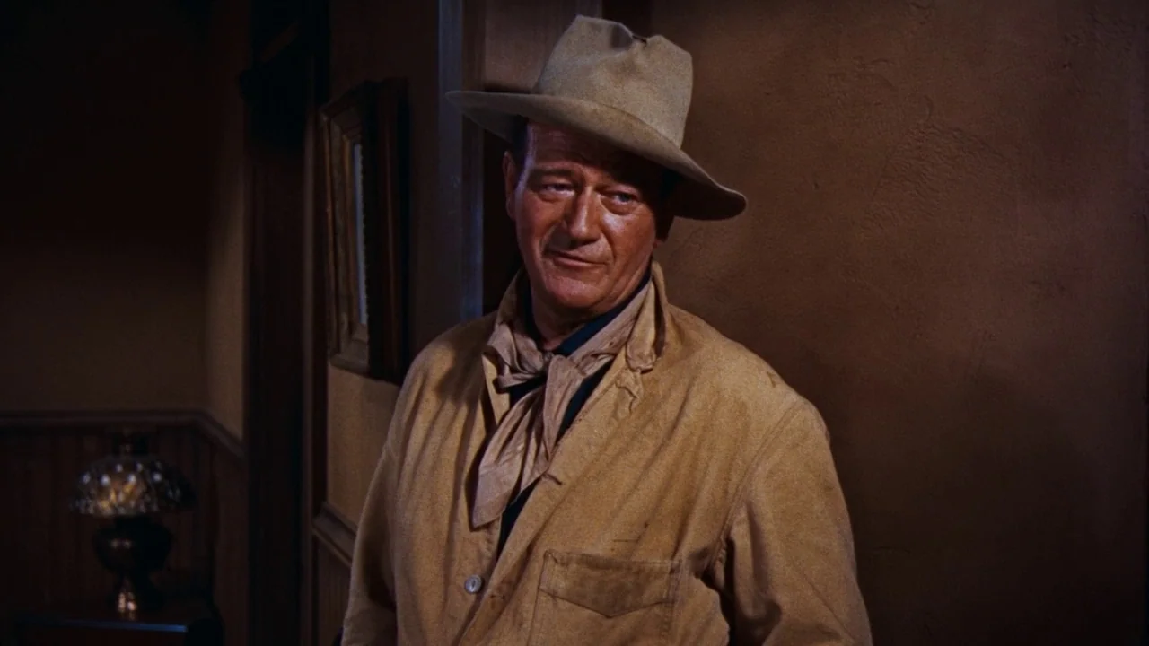 Rio Bravo (1959) movie still: medium — A dimly lit room with brown walls features a small table lamp. A man stands leaning against a wall while…