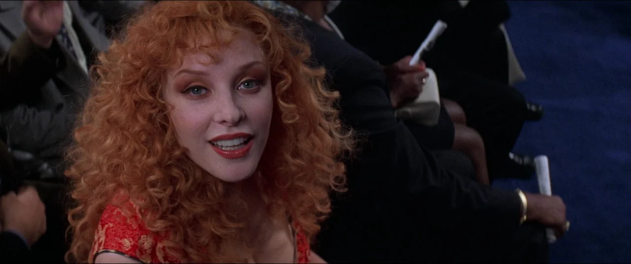 Snake Eyes (1998) movie still: closeup — A woman sits in a dark blue auditorium among other audience members. The camera focuses on her face as she…