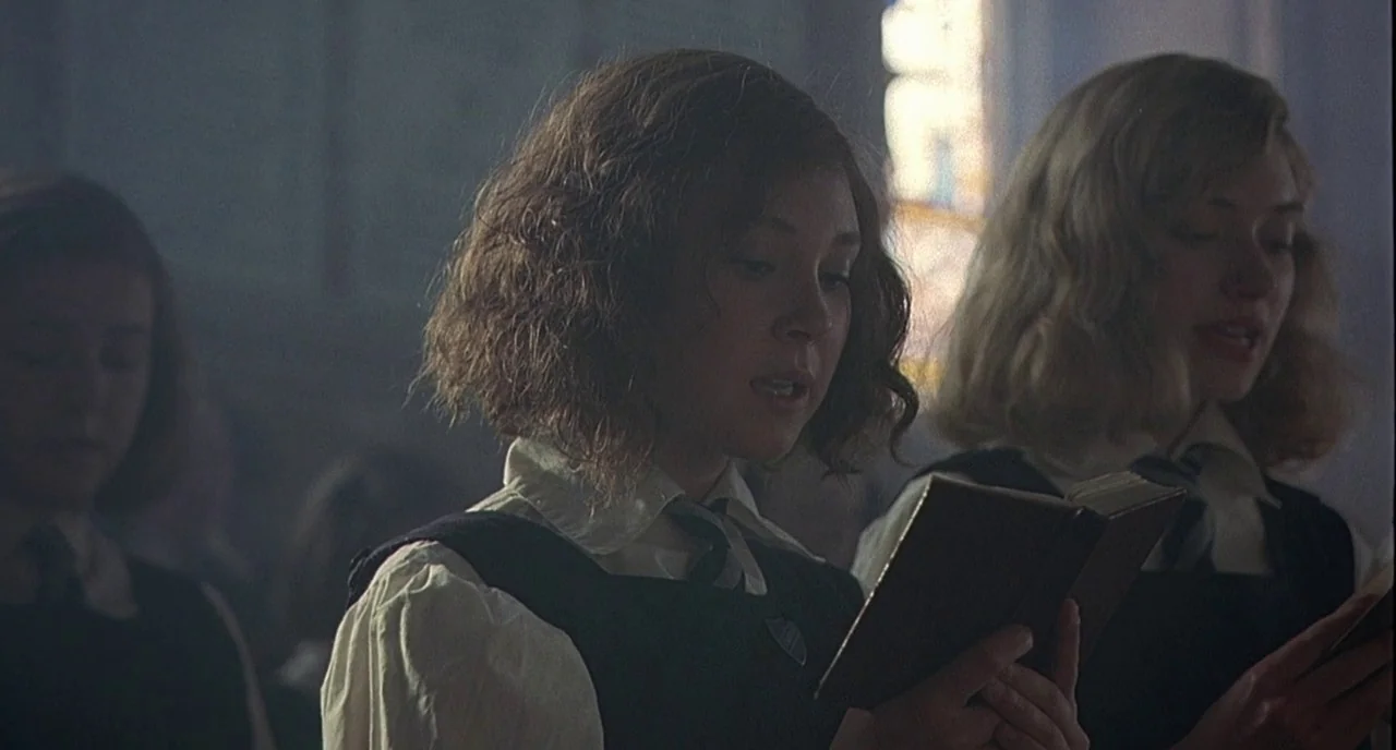Cracks (2009) movie still: medium — A group of schoolgirls in uniforms stand in a dim, light-filled religious space. The girls read from open books…