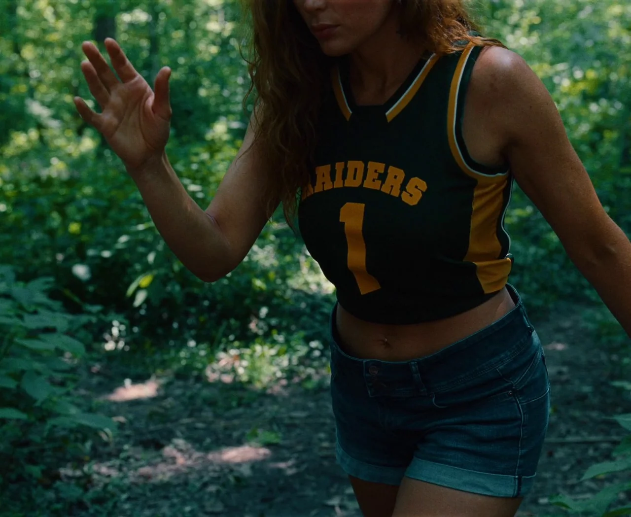 Monica (2022) movie still: medium — A woman walks through a dense, green wooded area during the day. She wears a athletic tank top and denim shorts…
