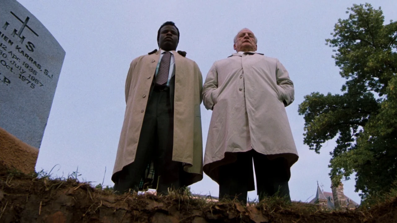 The Exorcist 3 (1990) movie still: fullbody — Two men stand together in an overcast cemetery looking down into a freshly dug grave. A grey stone…