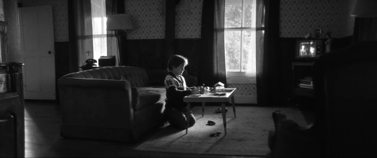 The Eyes of My Mother (2016) movie still: wide — A dark living room features floral wallpaper and a television set playing in the corner. A young…