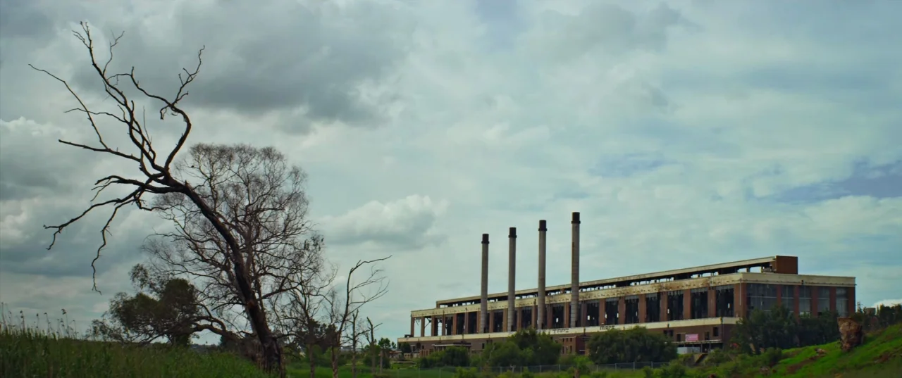 Chappie (2015) movie still: establishing — A sprawling industrial facility with four tall smokestacks sits under a cloudy sky. In the foreground, a…