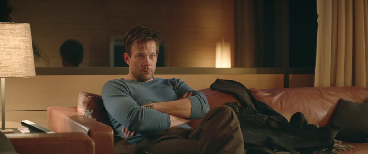 Force Majeure (2014) movie still: medium — A dimly lit room features a leather sofa and a lamp in the foreground. A man with crossed arms sits on…
