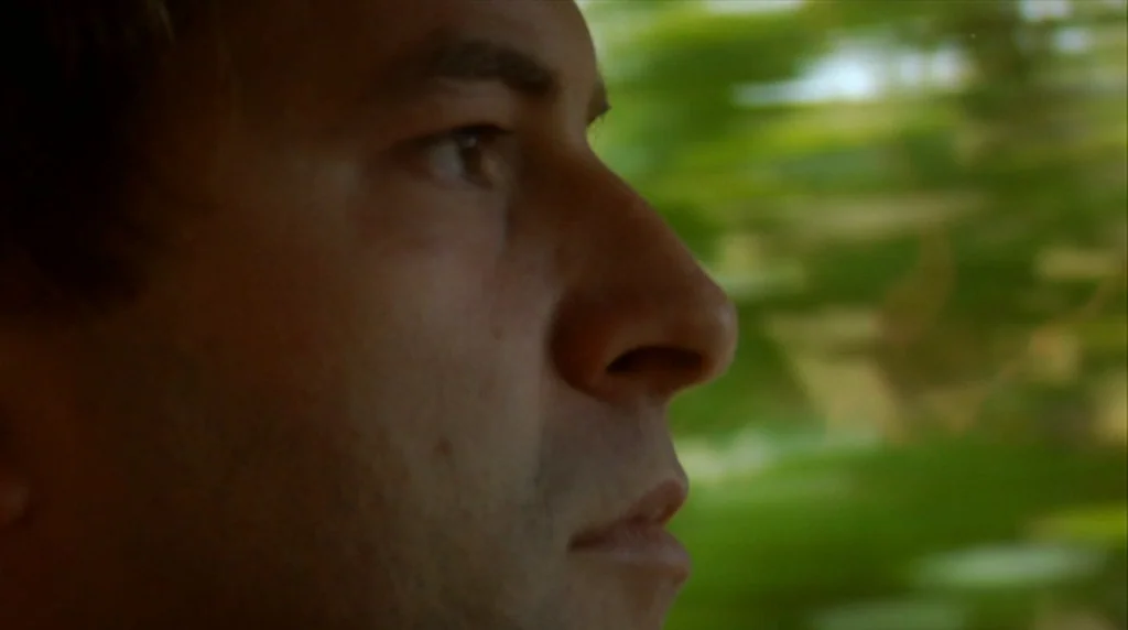 Humpday (2009) movie still: closeup — A side profile of a man seen from inside a moving vehicle. Green foliage blurs past the window in the…