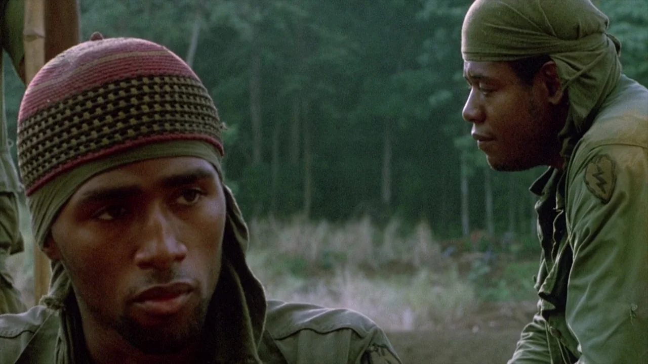 Platoon (1986) movie still: medium — Two men wearing military field gear are positioned in a lush green jungle setting. They appear to be talking or…