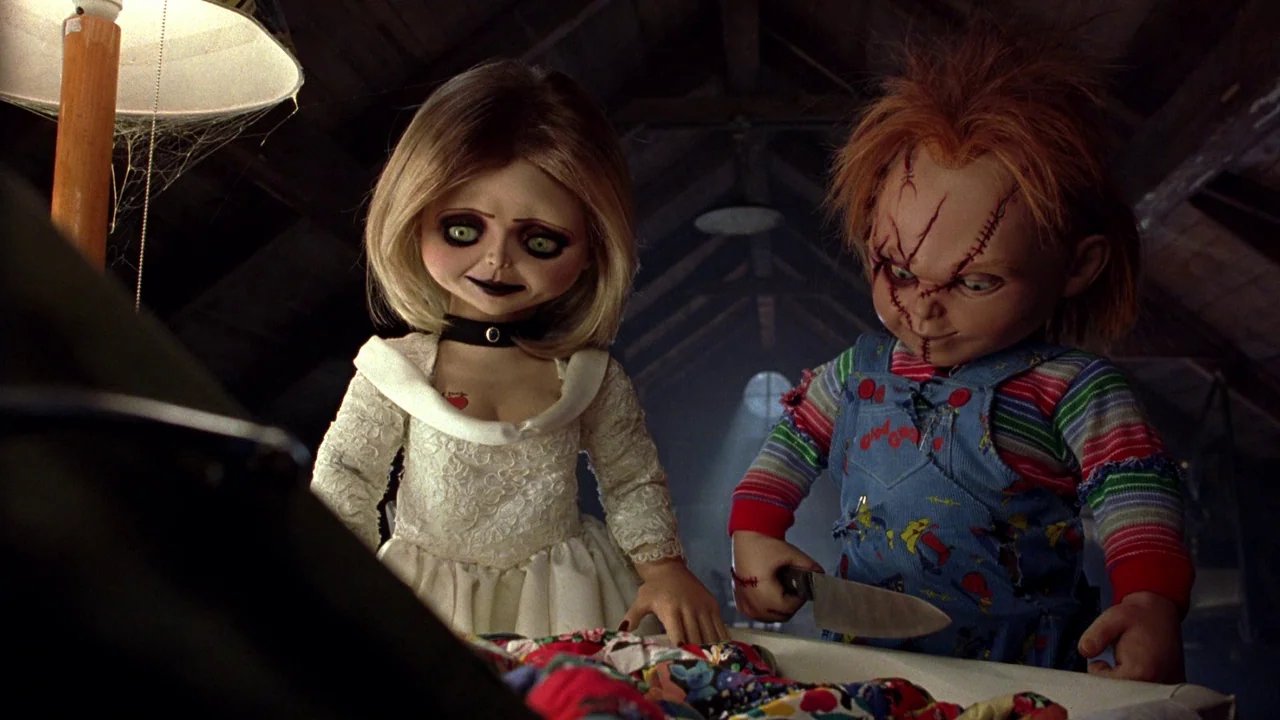 Seed of Chucky (2004) movie still: medium — Two small plastic dolls stand in a cluttered, dimly lit wooden attic. The male doll holds a large…