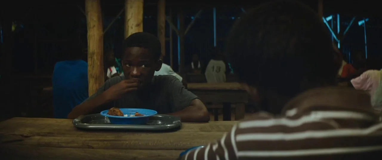 Beasts of No Nation (2015) movie still: medium — A dimly lit communal dining area features long wooden tables and benches for many people. A young…