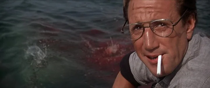 Jaws movie still: closeup — A man looks towards the camera from the side of a boat on water. Red blood creates a disturbance in the dark blue ocean…
