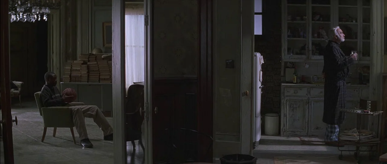 Finding Forrester (2000) movie still: wide — A dimly lit multi-room apartment contains stacks of books and traditional wood cabinetry. A younger man…