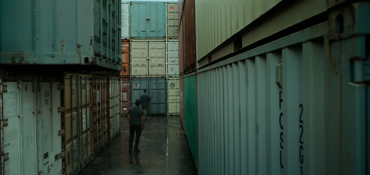 Triple Frontier (2019) movie still: wide — A narrow alley between towering stacks of rusted, colorful shipping containers in an industrial yard. Two…