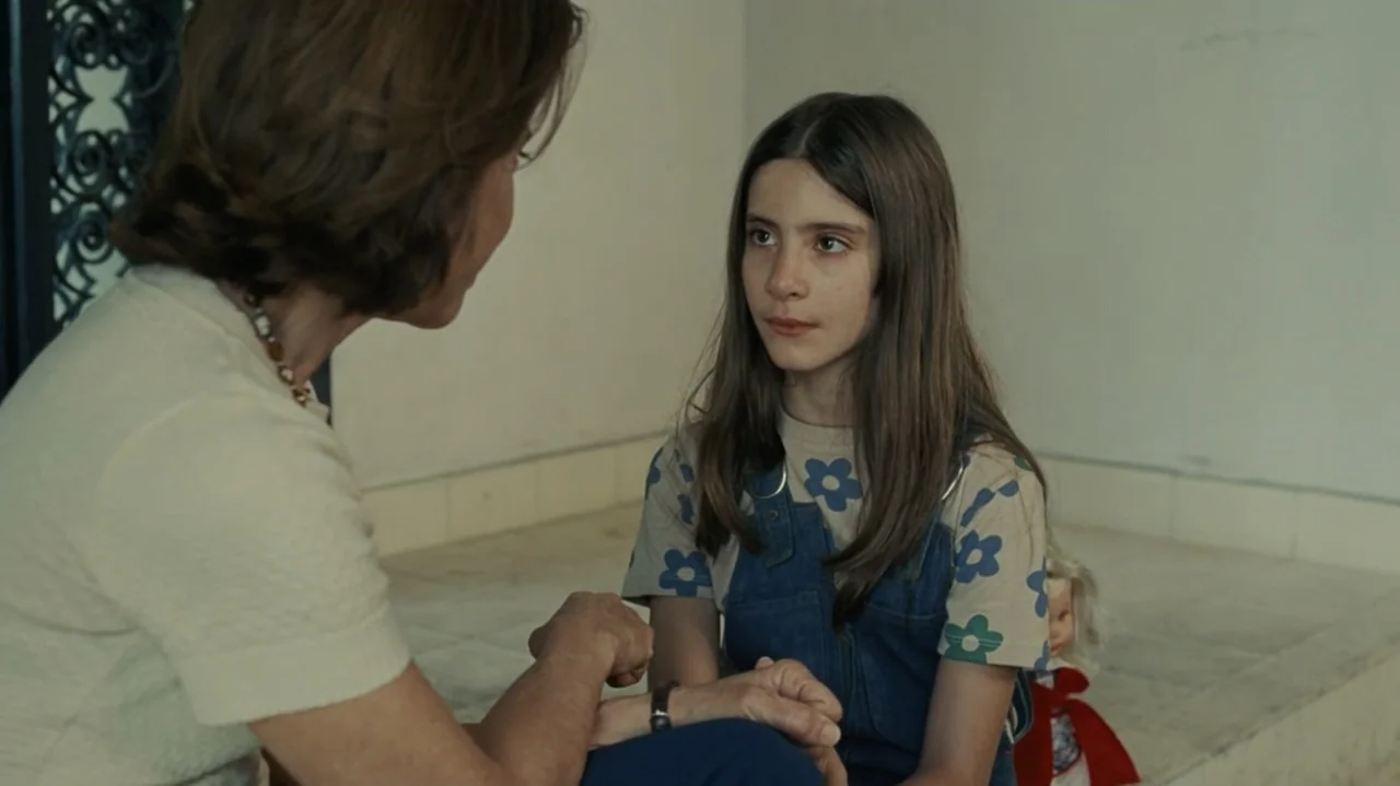 I'm Still Here (2024) movie still: medium — A plain interior room with light-colored walls and a marble-like floor. An adult woman sits facing a…