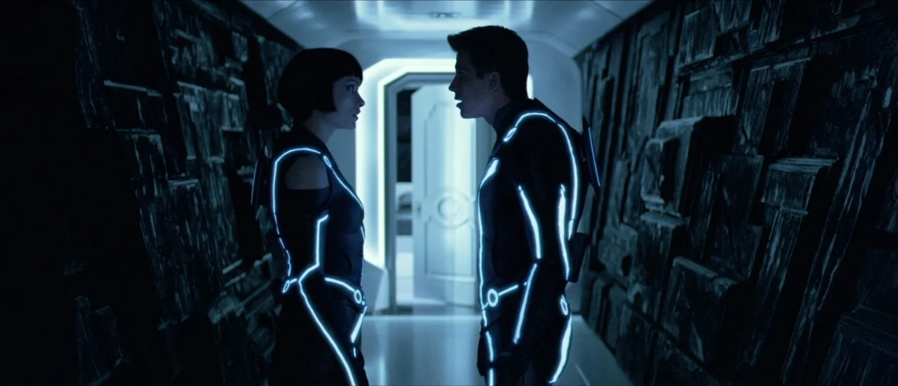 TRON: Legacy (2010) movie still: medium — A dark, textured hallway with geometric wall panels leads toward a bright white doorway. A man and woman…