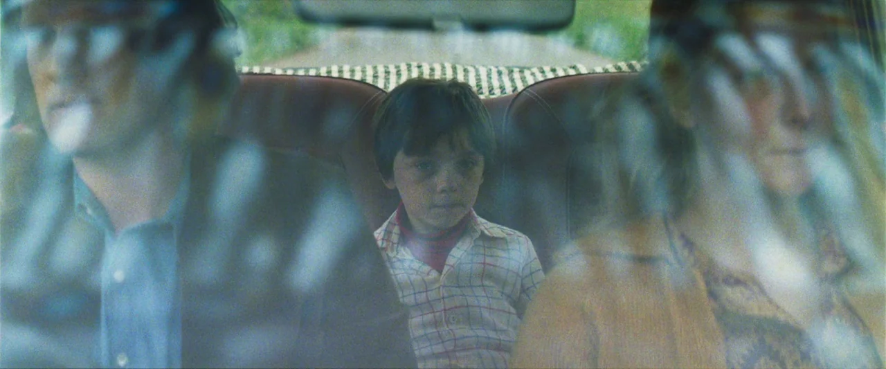 Starve Acre (2023) movie still: medium — Three people sit in the back seat of a vehicle with dark upholstery. A young boy centered in the frame sits…
