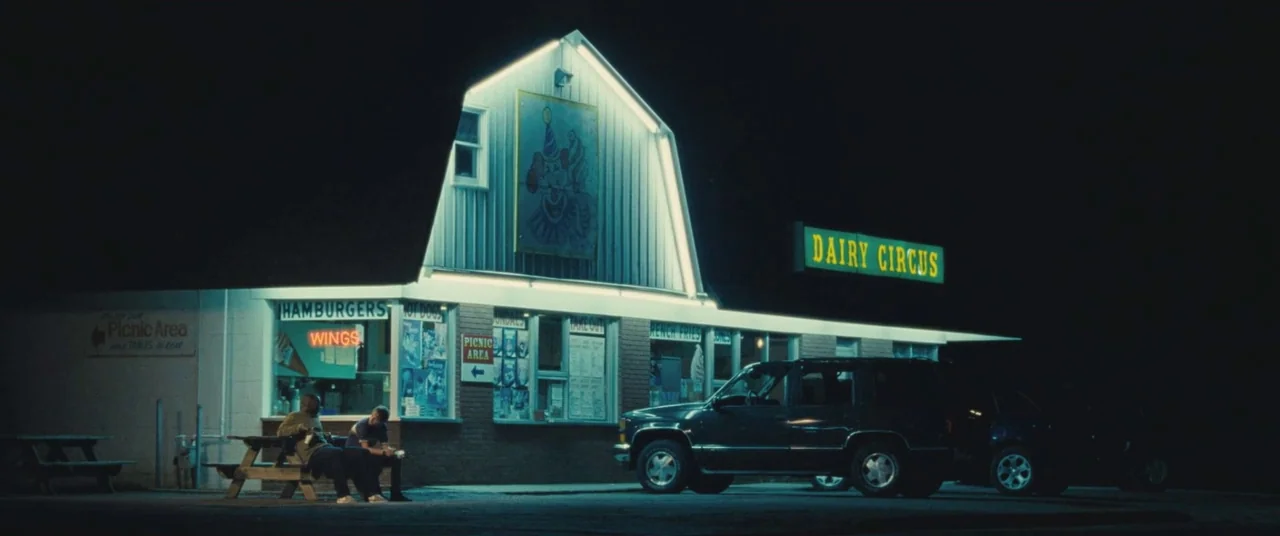 The Place Beyond The Pines (2012) movie still: wide — A night view of a small shop with barn-style architecture and illuminated neon signs. Two men…