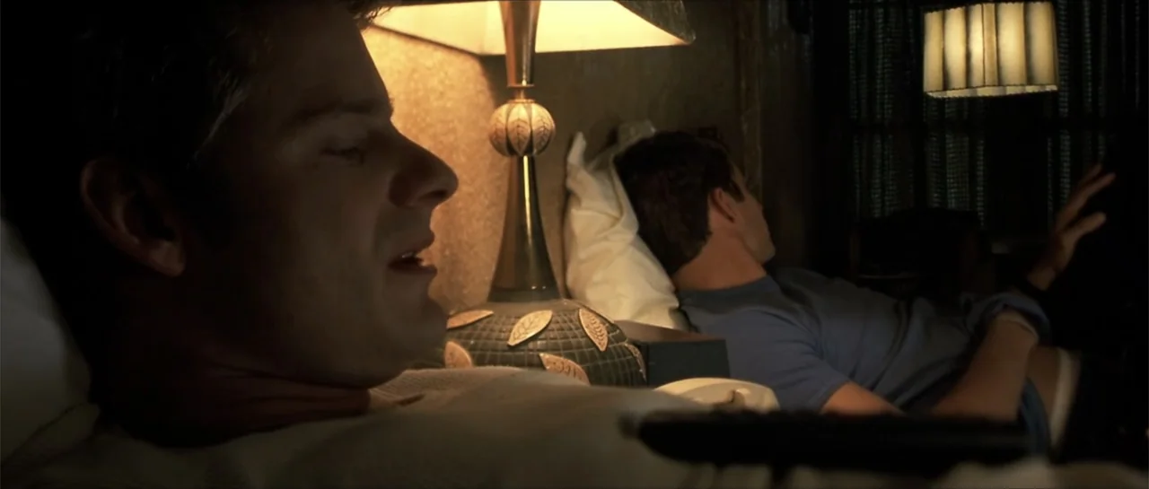 Joy Ride (2001) movie still: medium — A dimly lit motel room features a table lamp positioned between two bed pillows. Two men lie together in the…